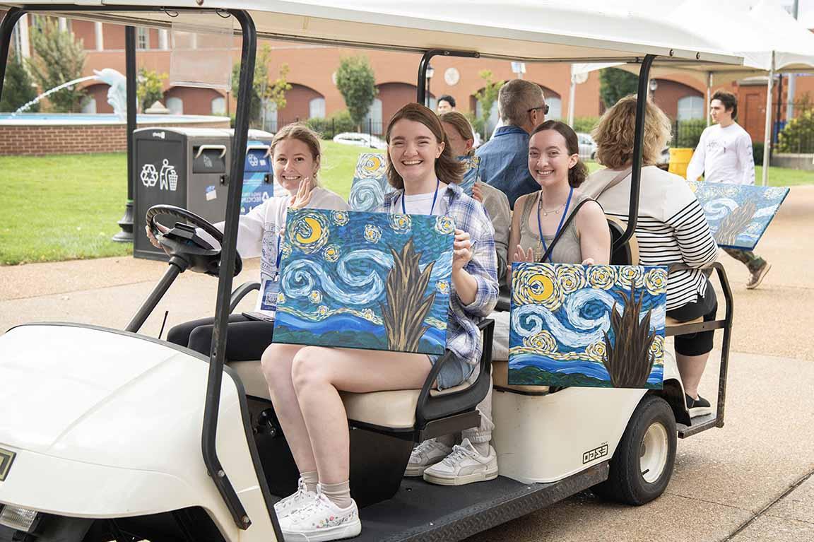 A golf car holding family members showing off artwork from the 2024 Homecoming and Family Weekend Paint and Sip drives down West Pine Mall. 