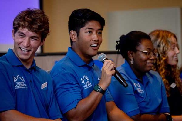 A student speaks into a microphone. Two students are seated next to him, smiling. 