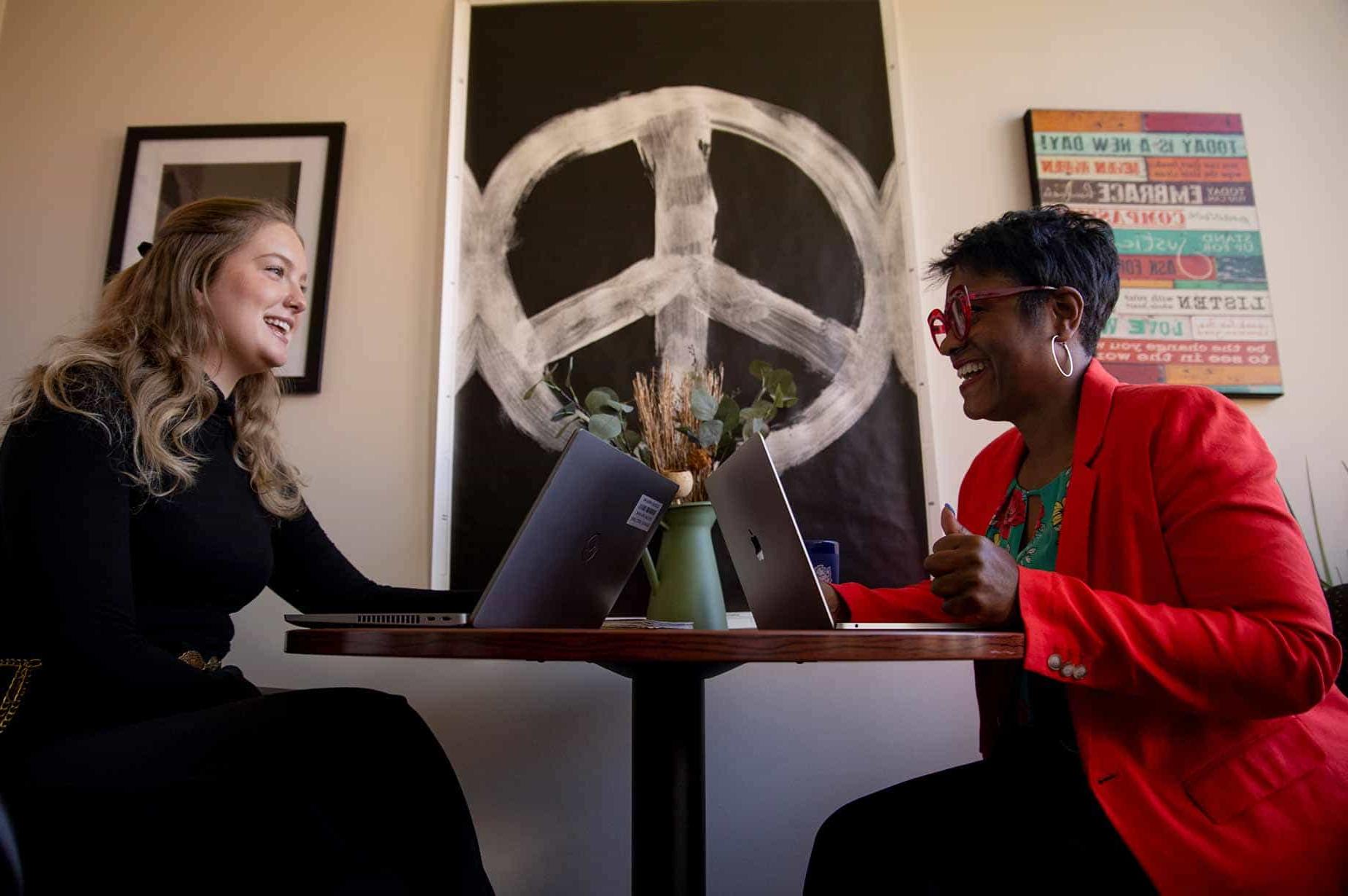 A female professor works with a group of students while sitting at a table. They are looking over paperwork and chatting.