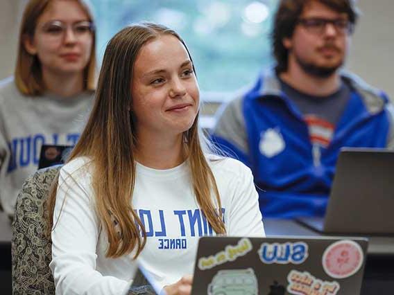 A student wearing a 博彩网址大全 Dance Shirt with a laptop covered in stickers looks attentively toward the front of a classroom