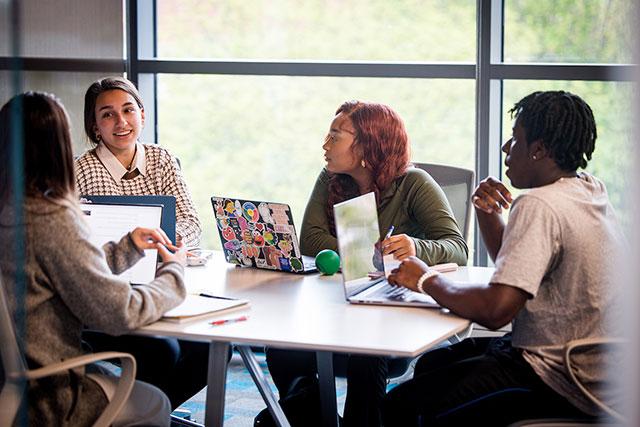 Four students with laptops sit at a table talking. 