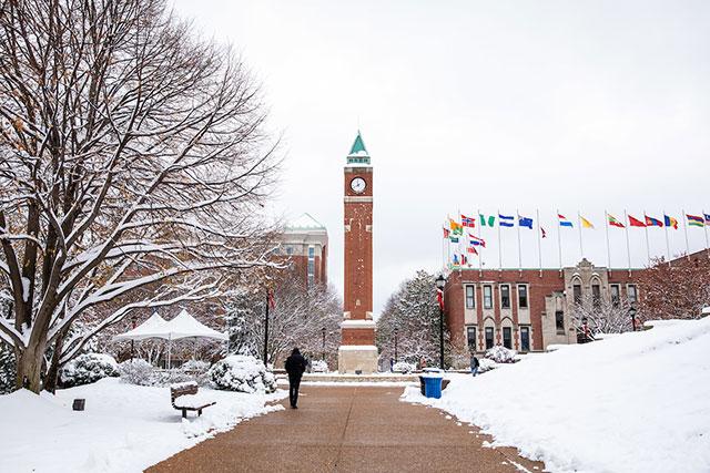 A person is seen from behind as they walk along a snowy campus toward the clock tower and center for global citizenship