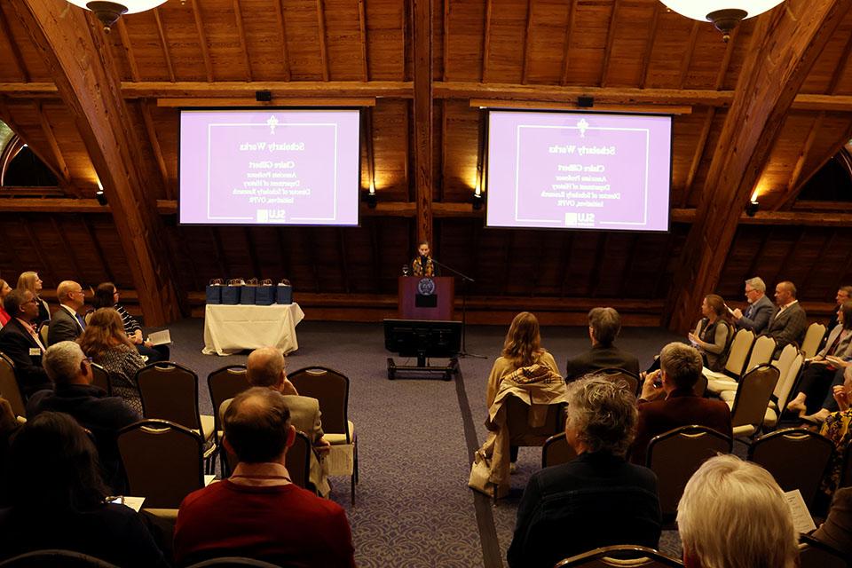 A woman stands at a lectern in front of an audience, with a video screen on each side