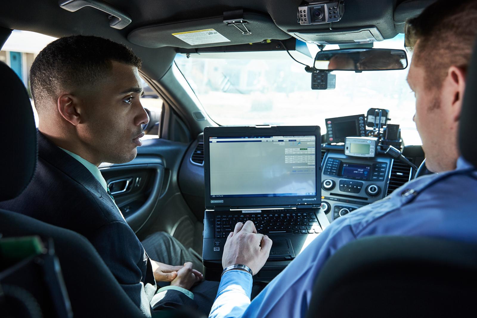 A student and a law enforcement officer look at a computer screen while sitting in a police car.